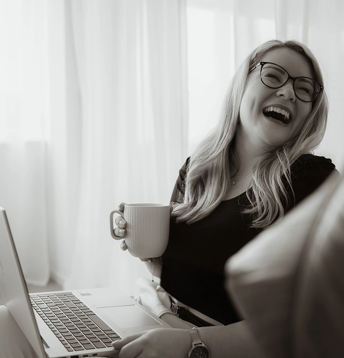 Berthilde Bittner sitzt entspannt mit Laptop auf dem Schoß und Kaffeetasse in der Hand, lacht herzlich in einem hellen Raum mit weißen Vorhängen – Beblick Business Coaching Regensburg für Unternehmer und Führungskräfte.